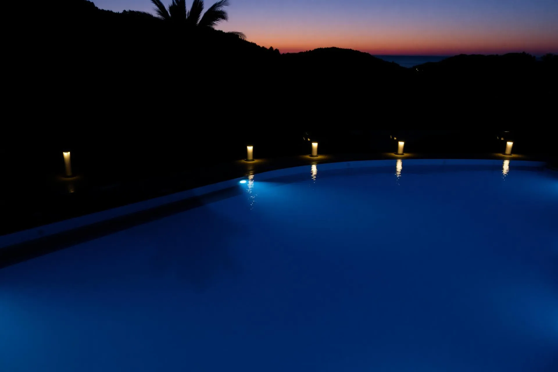 Piscina iluminada con agua azul al atardecer y palmeras al fondo